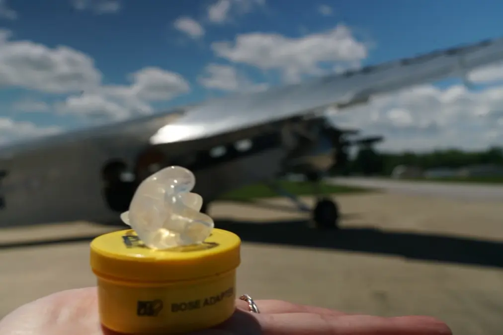 A clear custom Bose ProFlight adapter on its Big Ear case with a vintage aircraft behind